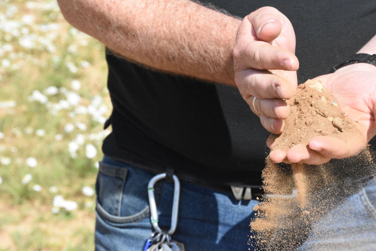 A view of someone holding sandy soil in their hands as it trickles their fingers. They're wearing jeans with a karibiner attached to one of the belt loops.