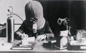 A black and white photograph, apparently from the early 20th century, showing a person sitting at a desk writing, wearing a large padded helmet that completely encases their head. The helmet has small goggles over the eyes and a breathing apparatus over the mouth, connected by a tube to a gas cylinder on the left. A desk clock, telephone, and other office items are visible on the desk.