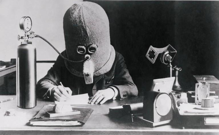 A black and white photograph, apparently from the early 20th century, showing a person sitting at a desk writing, wearing a large padded helmet that completely encases their head. The helmet has small goggles over the eyes and a breathing apparatus over the mouth, connected by a tube to a gas cylinder on the left. A desk clock, telephone, and other office items are visible on the desk.