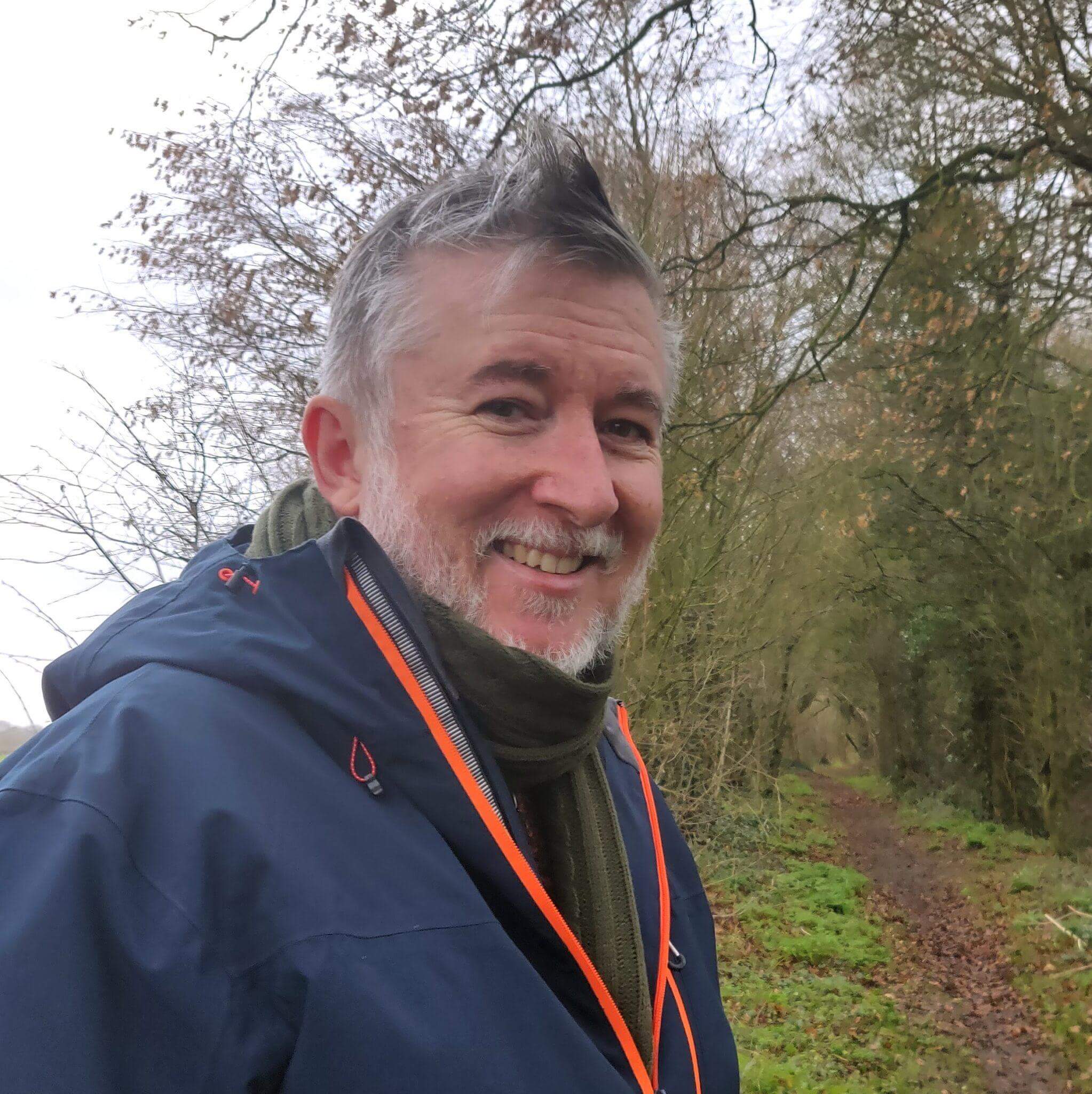 Nathan Nelson smiling outdoors on a woodland path, wearing a blue jacket with orange zip