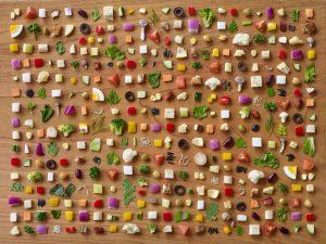 Hundreds of small chopped salad ingredients arranged in a grid on a wooden surface, including vegetables, olives, eggs, and cheese